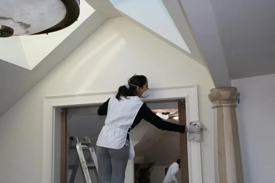  Person on ladder cleaning a high ceiling corner in a room, wearing a mask and gloves.