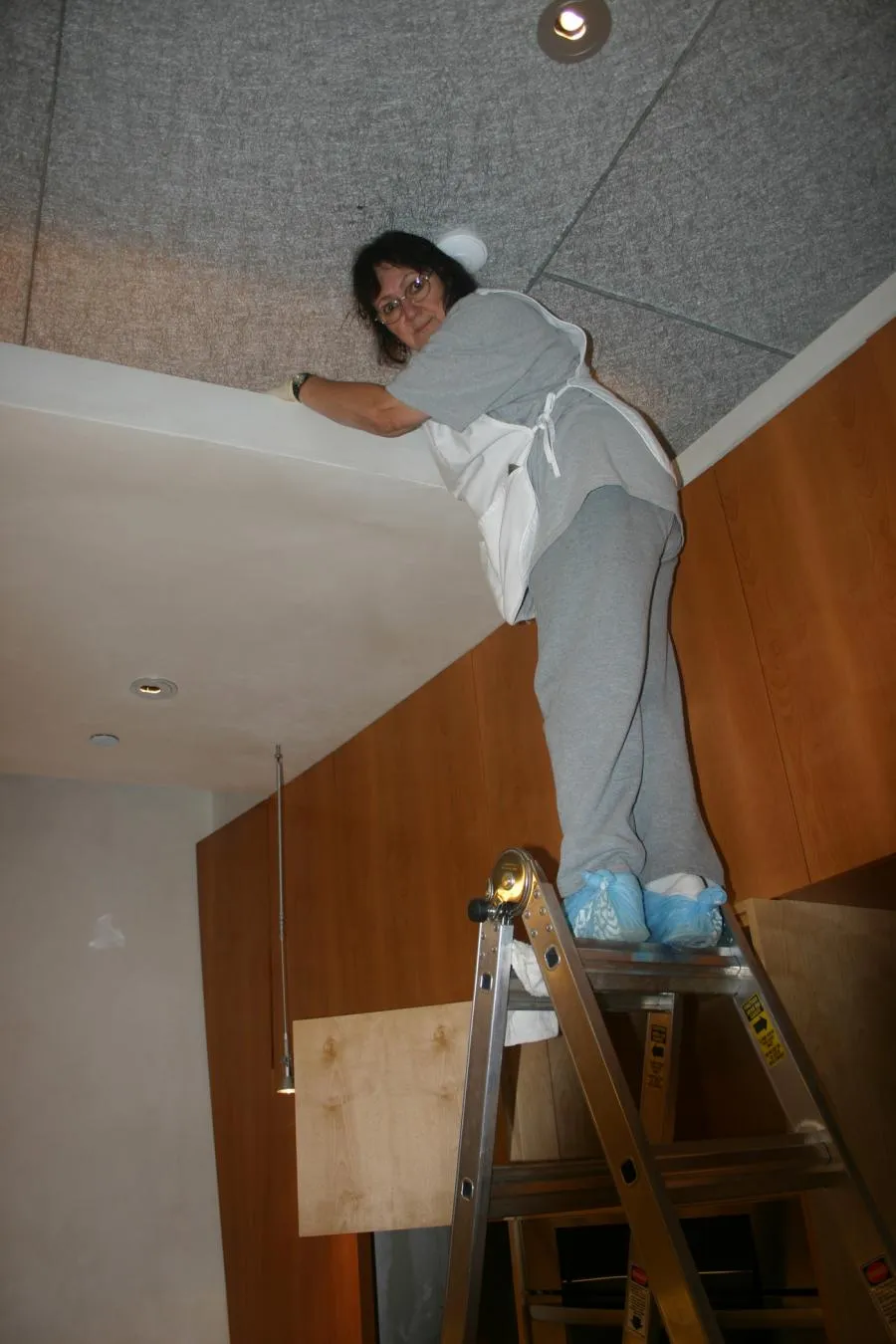  Person standing on a ladder cleaning a ceiling, wearing grey clothes and blue shoe covers, inside a room with wooden cabinets.