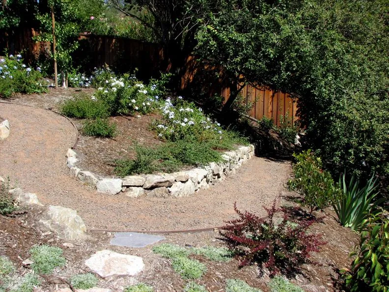  Curved gravel path bordered by stones, surrounded by green shrubs and flowers under clear daylight.
