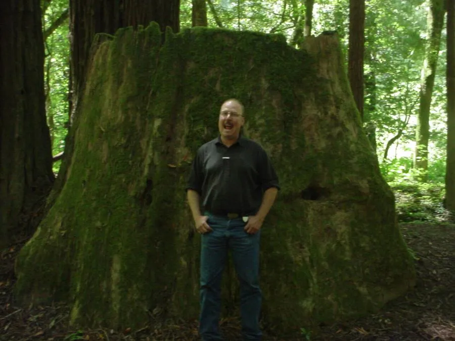  A man stands smiling in front of a large, moss-covered tree stump in a forest.