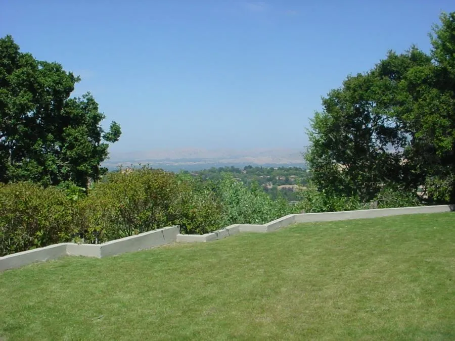 Manicured lawn with concrete edging, surrounded by shrubs and trees, overlooks a distant landscape under a clear blue sky.