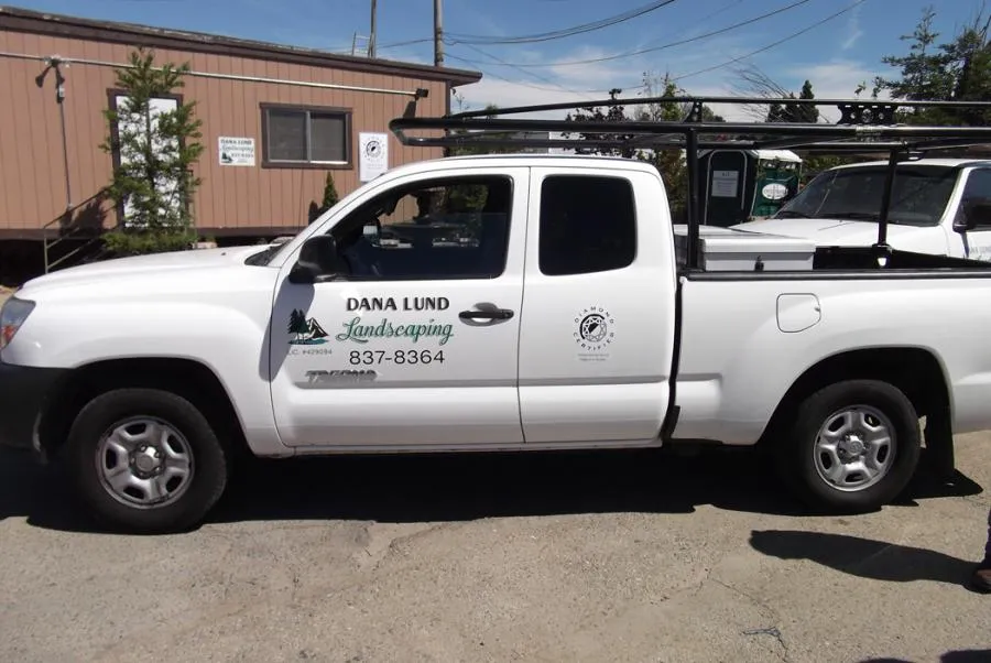  White Dana Lund Landscaping truck parked in front of a building with visible company logo and contact information.