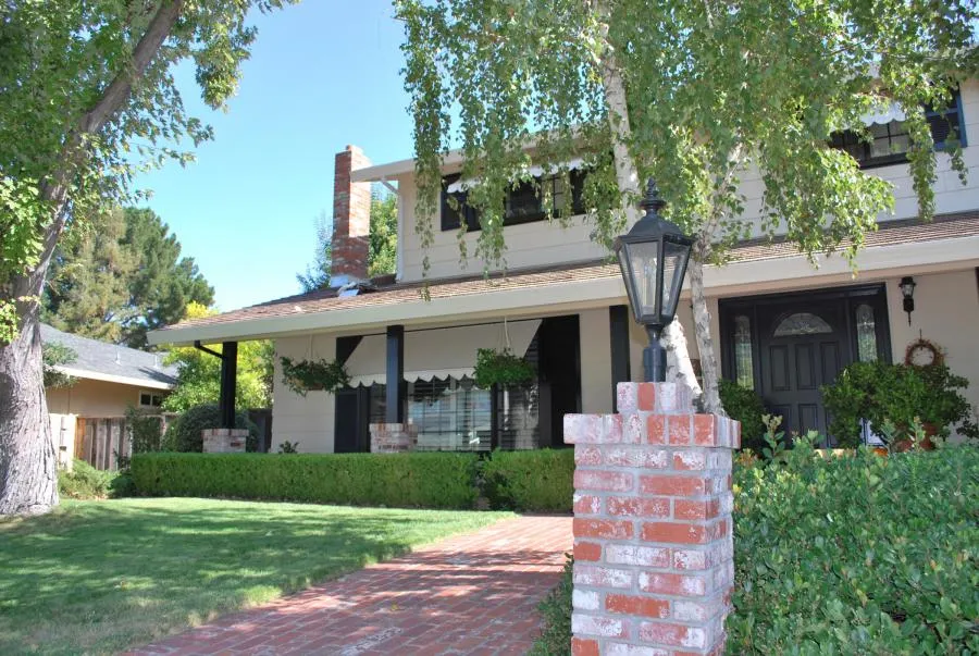  Front view of a two-story house with a brick pathway, lush lawn, and large trees. The home's exterior features a porch and classic black door.