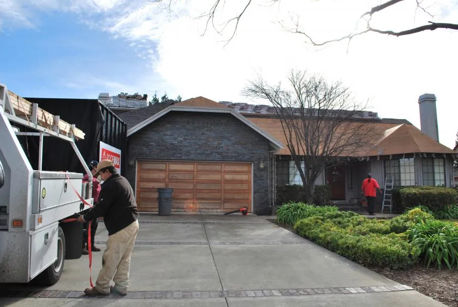 A worker stands by a truck with roofing materials at a house with unfinished roofing, another worker near a ladder, and garden in front.