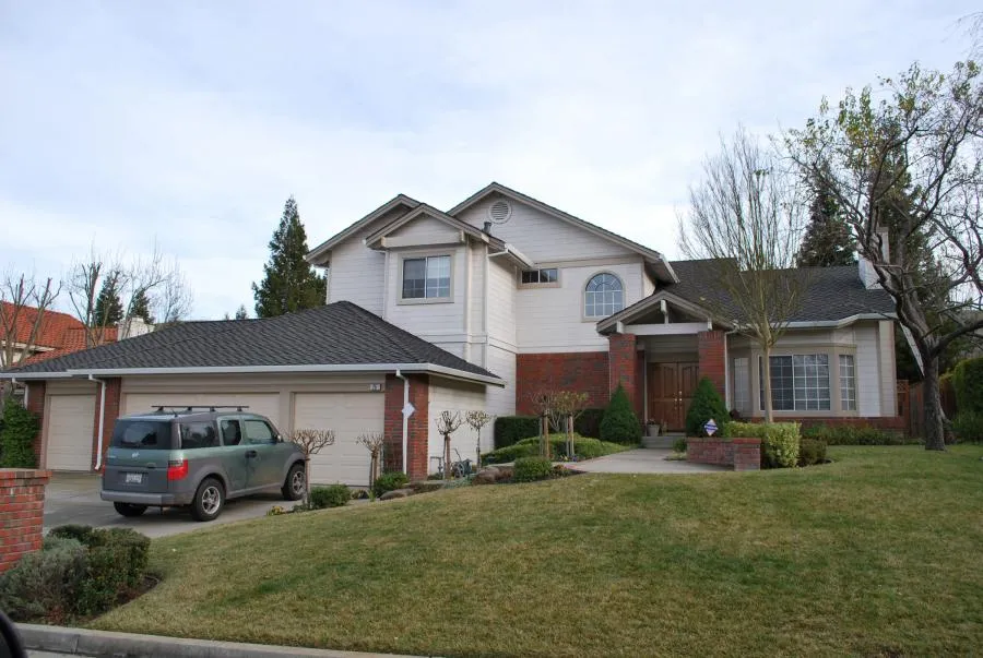 A two-story suburban house with a dark shingle roof, brick facade, and a manicured front lawn. A large tree and bushes frame the entryway. An SUV is parked in the driveway.