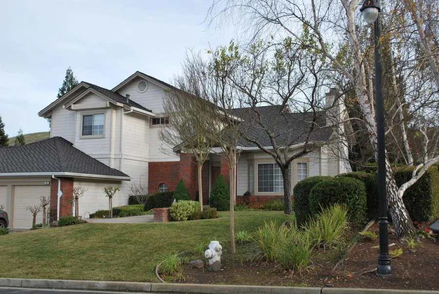  A suburban house with a dark shingle roof, light siding, brick accents, and manicured landscaping under a cloudy sky.