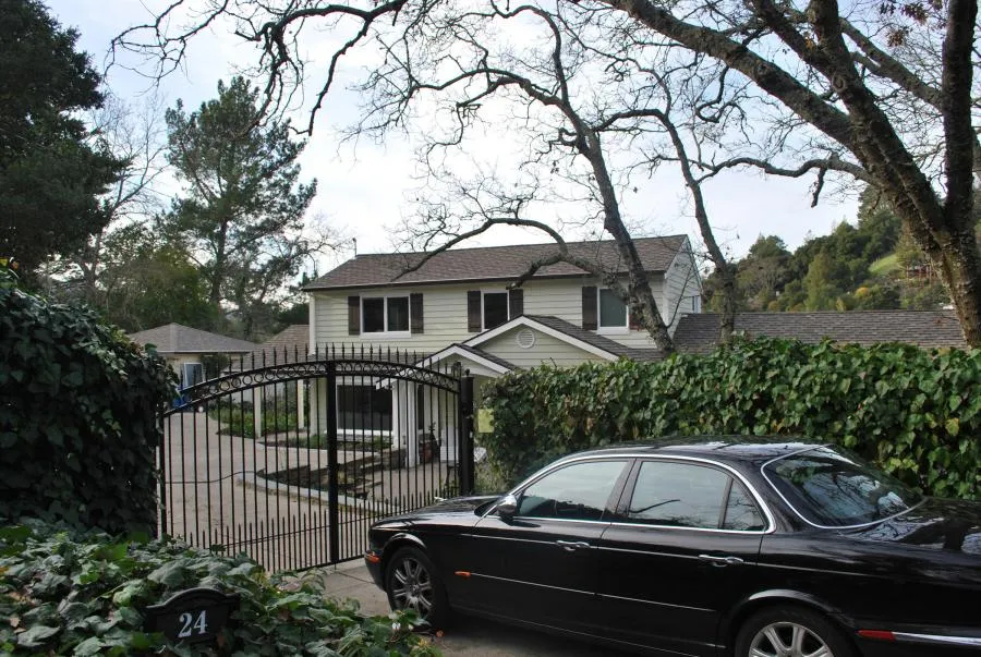 Suburban house with a black car parked in front of a closed iron gate, surrounded by trees and ivy-covered walls.