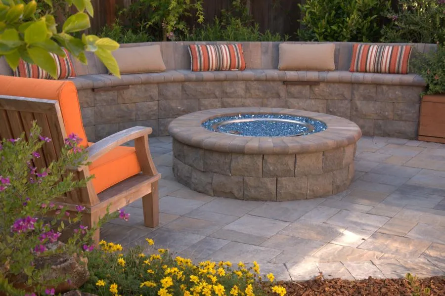 Picture of A serene outdoor seating area by Black Diamond Paver Stones & Landscape, Inc. features a circular stone fire pit surrounded by a curved stone bench with colorful cushions. Renowned for focusing on quality outcomes, the company creates inviting spaces for relaxation and gathering. Copyright ©2025 Diamond Certified Resource. - Black Diamond Paver Stones & Landscape, Inc.