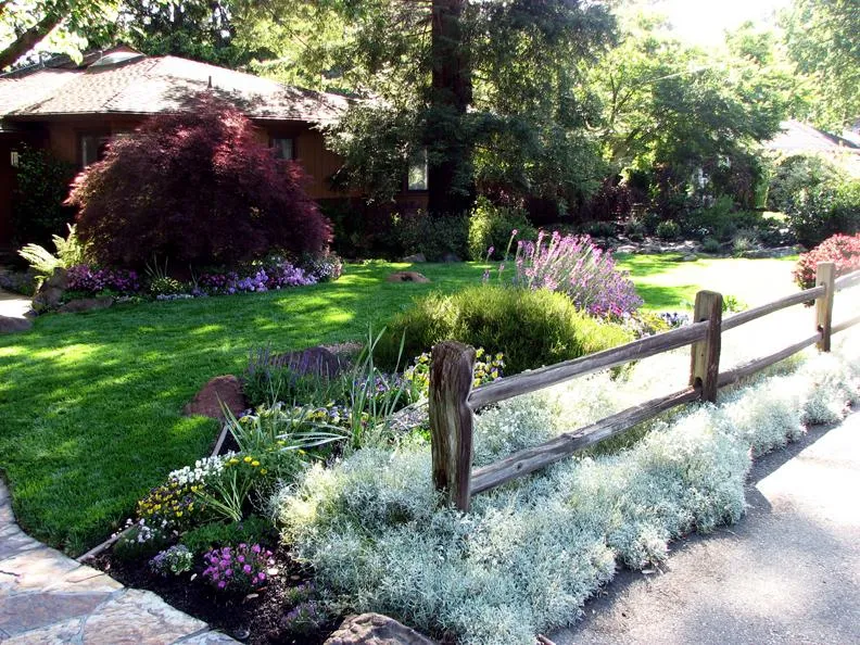 A landscaped garden with green lawn, diverse plants, a rustic wooden fence, and a stone path leading to a house.