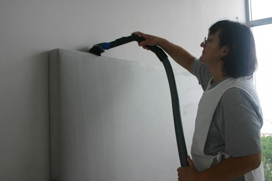  Person cleaning an upholstered wall panel with a vacuum cleaner attachment in a bright room.