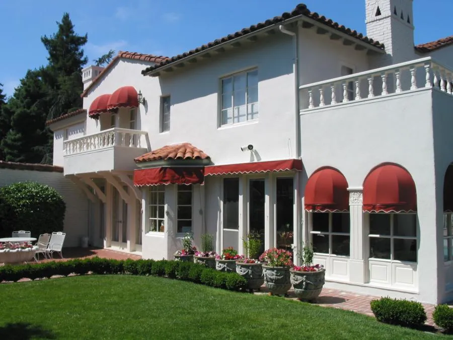  A white house with red awnings over windows and doors, a balcony, and manicured lawn with shrubs.
