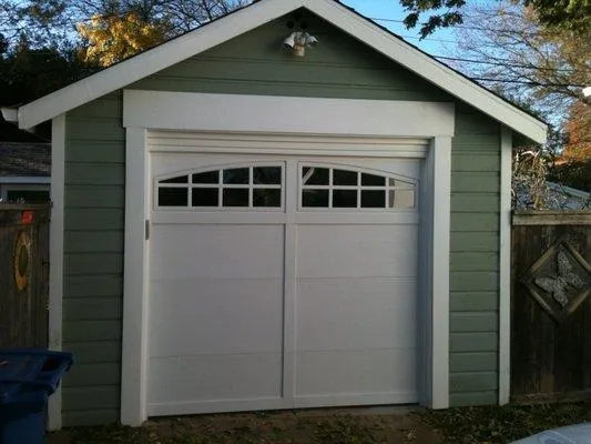  White garage door with arched windows installed on a green-painted garage, surrounded by a wooden fence and trees in the background.