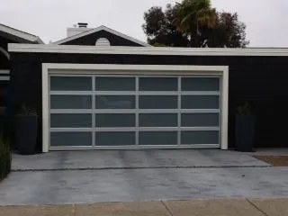  Modern garage door with frosted glass panels, framed by a dark exterior wall and white trim, flanked by tall potted plants.