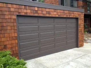  A modern garage door with a brown finish is set in a building with red cedar shingle siding. The concrete driveway leads up to the garage entrance.