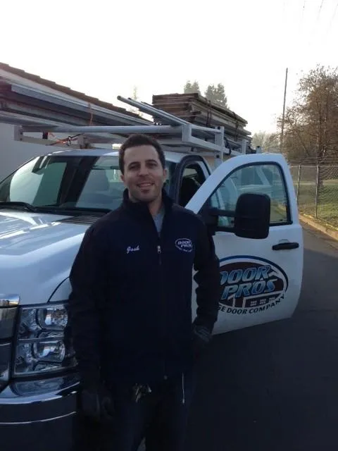  Man in a Door Pros, Inc. jacket stands next to a company truck with ladders on top, in an outdoor setting.