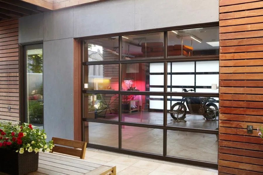 Modern glass garage door with a view into an interior space containing a motorcycle and decor, surrounded by wooden paneling and patio furniture.