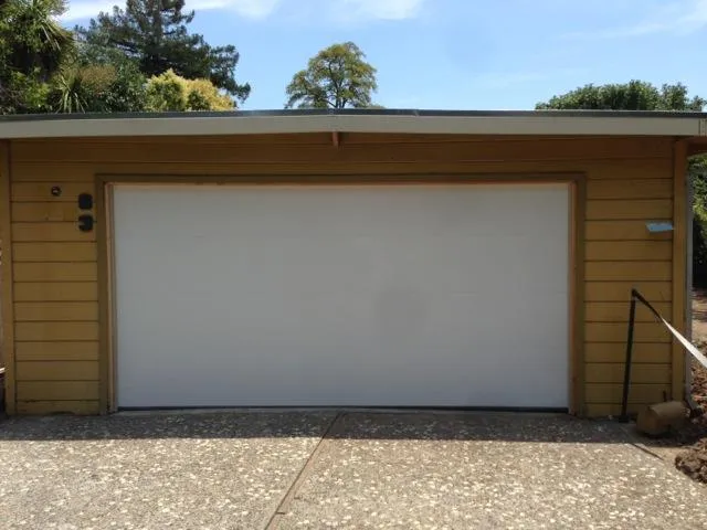 Closed white garage door set in a mustard yellow building with a concrete driveway in front. Trees and sky visible in the background.