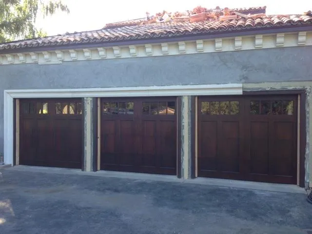  Three wooden garage doors installed in a row on a stucco building with a tiled roof.