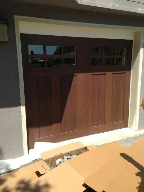  Wooden garage door with six upper glass panels, partially open, surrounded by white trim, with cardboard and hardware on the driveway.