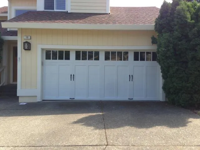  Newly installed white garage door with windows on a residential home, surrounded by a yellow exterior and greenery.