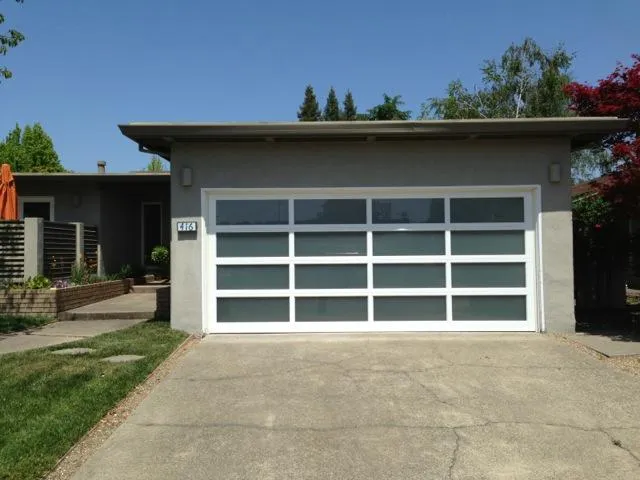  Modern glass-paneled garage door installed on a residential garage, surrounded by a concrete driveway and landscaped yard.
