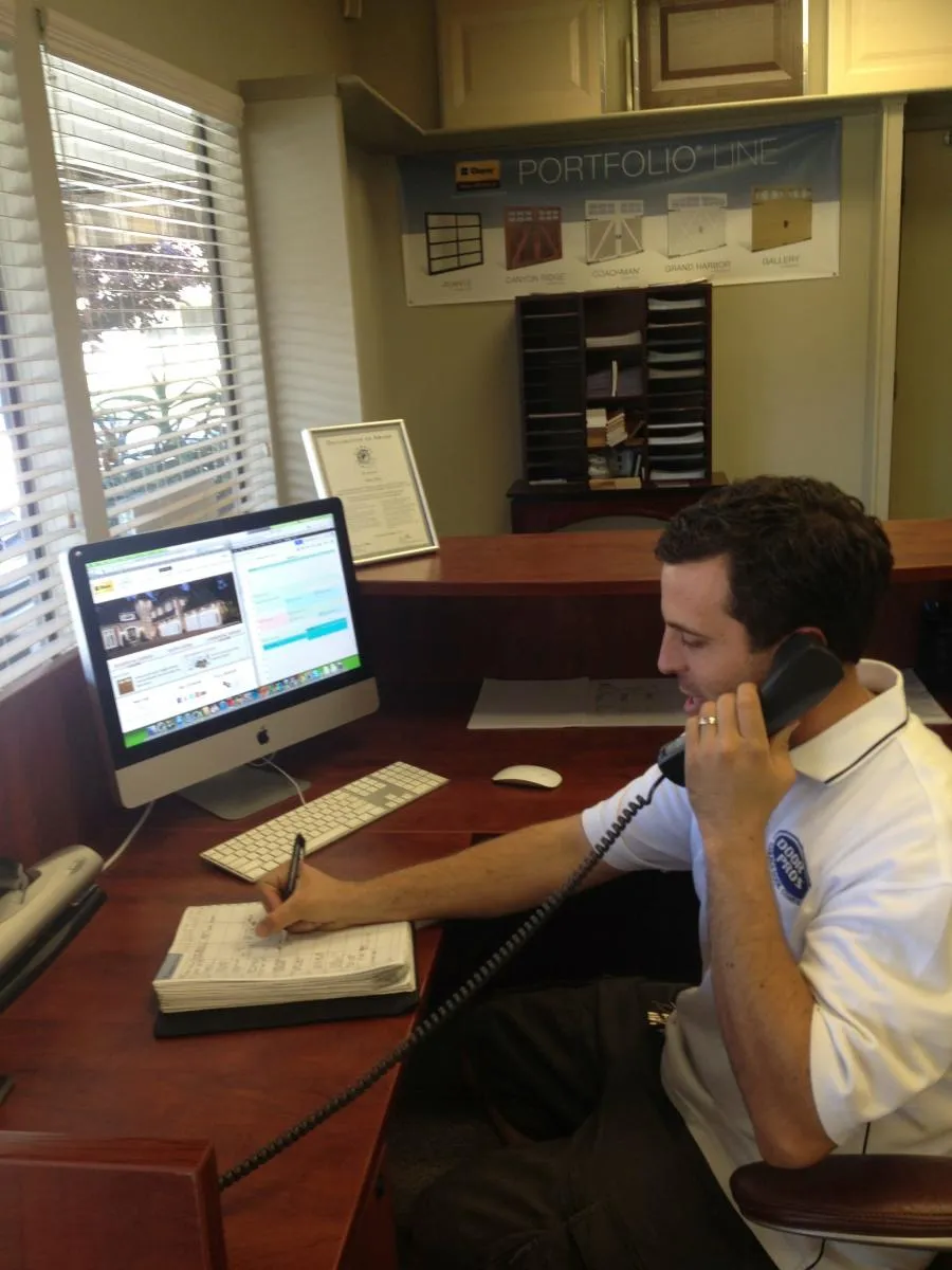  Person in uniform at desk using a phone and writing in a planner, with a computer and office supplies visible around.