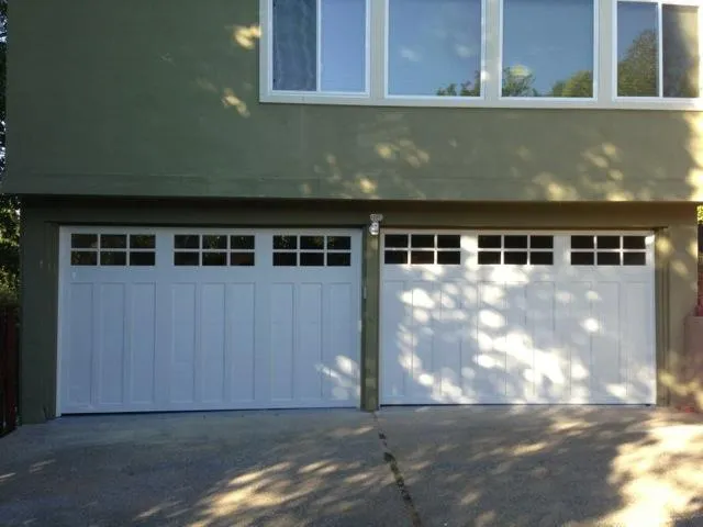  Two white garage doors with windows are installed under a building's shaded exterior.