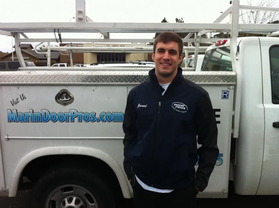 A man in a branded jacket stands in front of a Door Pros, Inc. service truck with visible company logo and website.
