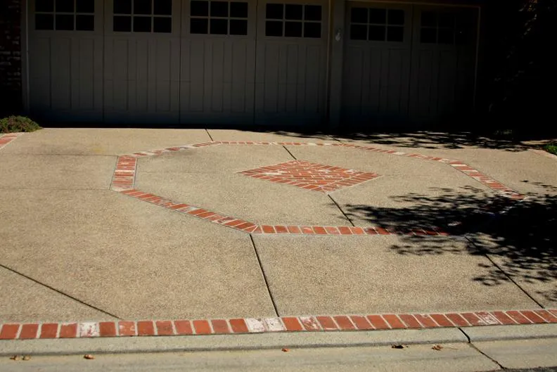Driveway with a geometric brickwork design, featuring a central square surrounded by brick outlines, leading to a garage with closed doors.
