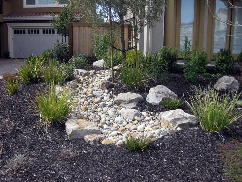  Dry creek bed with stones and surrounding plants in a landscaped garden in front of a house.
