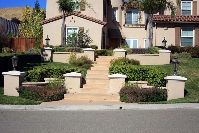  Front view of a house with beige stucco, featuring landscaped steps, shrubs, and decorative lighting on a well-maintained lawn.