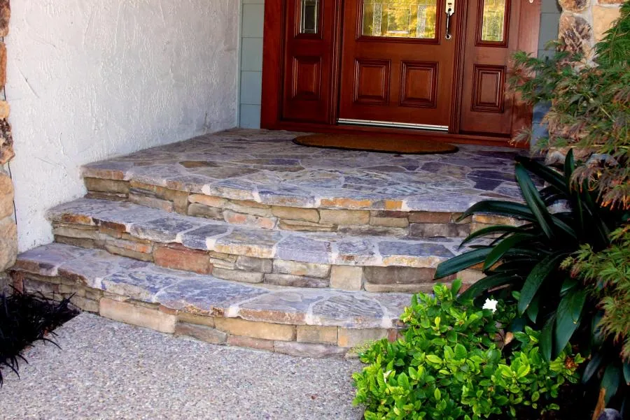  Stone steps leading to a wooden front door, surrounded by plants and greenery, showcasing detailed stonework and landscaping.