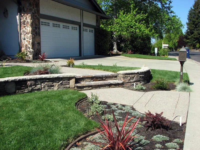 Stone retaining wall, manicured lawn, and pathway leading to a garage, surrounded by neatly arranged plants and greenery.