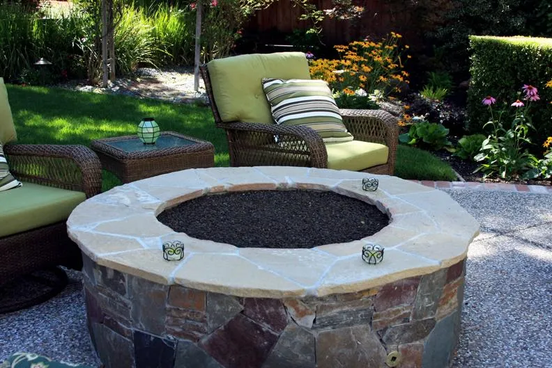  Stone fire pit surrounded by wicker chairs with green cushions in a landscaped garden. Lush grass, flowers, and shrubs in the background.