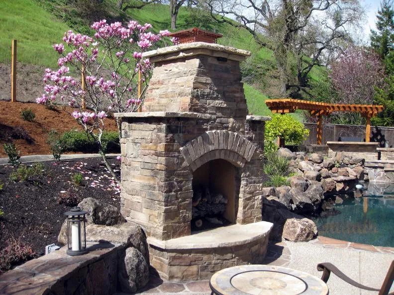  Stone fireplace on a landscaped patio with blooming pink flowers and a pool, surrounded by greenery and rock features.