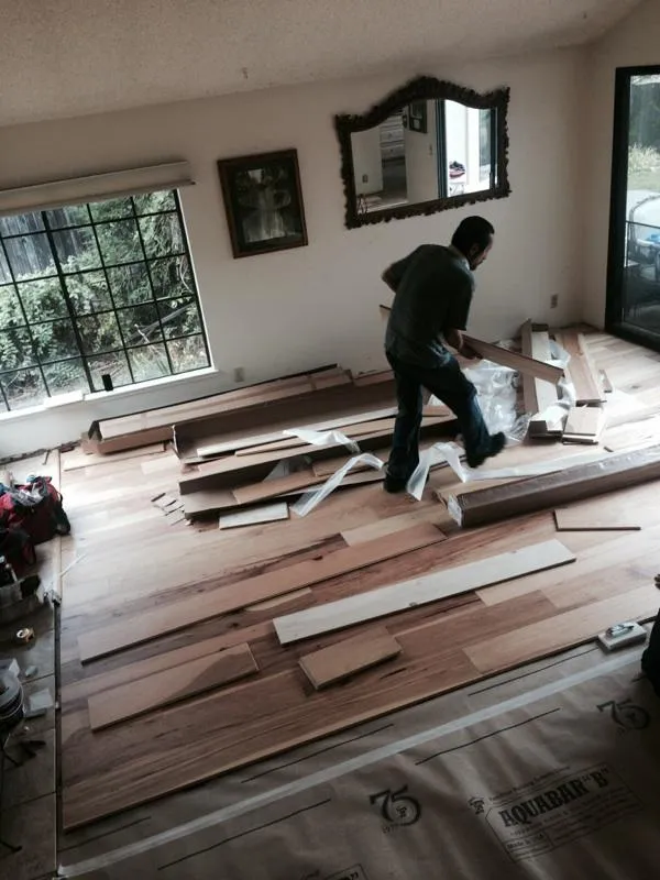  A worker lays wooden planks on a living room floor, surrounded by tools and materials. Large windows and a wall mirror are visible.