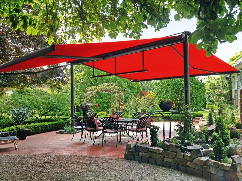 A red sunshade covers a patio with metal chairs and table surrounded by lush gardens and stone landscaping.