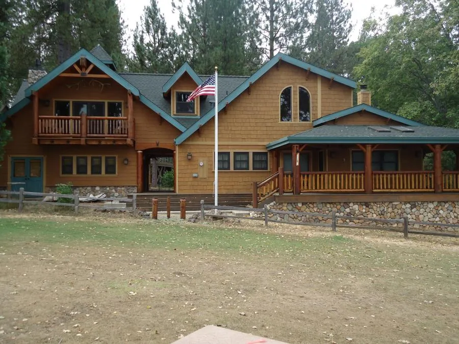  A large two-story house with a wooden exterior, stone foundation, and an American flagpole, surrounded by trees and a wooden fence.