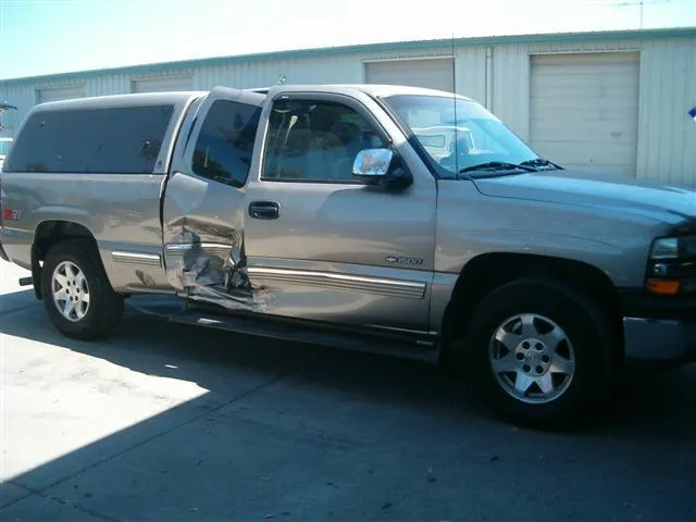  A silver truck with a damaged side door is parked in front of a garage, showing visible dents on the vehicle.