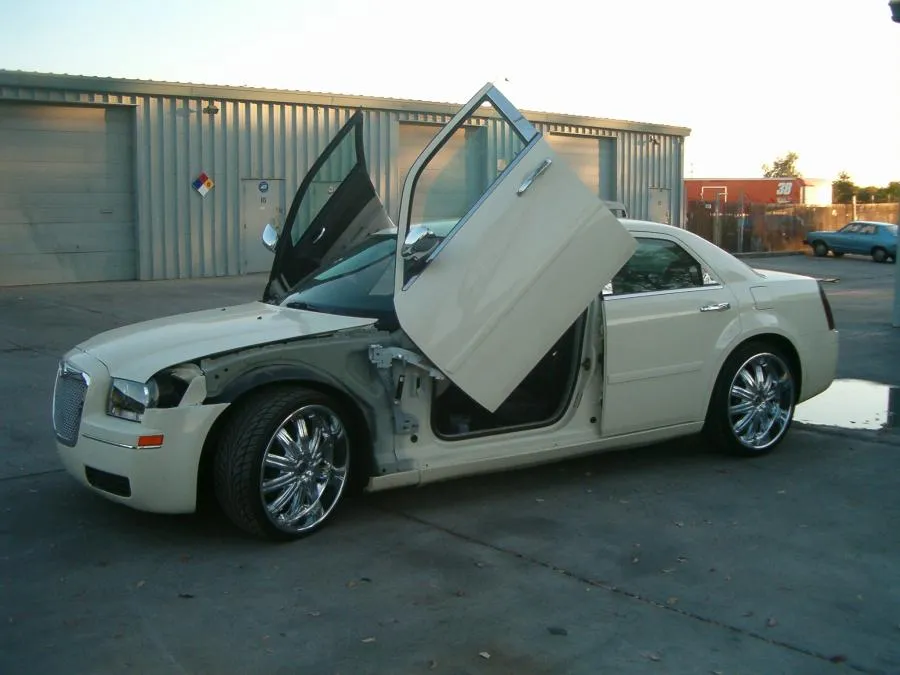 White car with open vertical doors and exposed front panel parked in a lot, showcasing custom modifications.