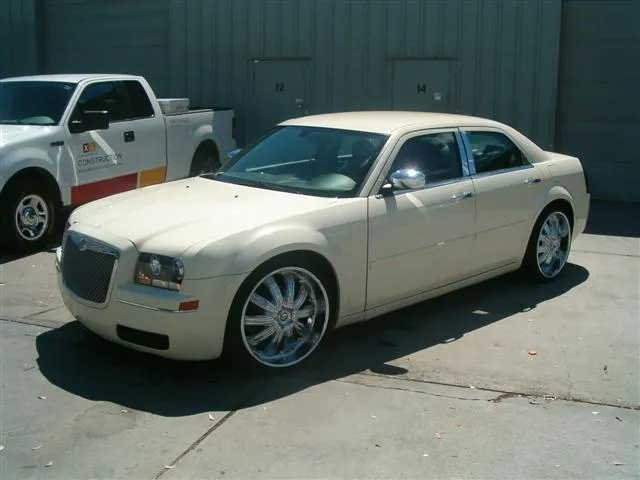 Cream-colored sedan with shiny chrome wheels parked in front of a gray building beside a white truck.