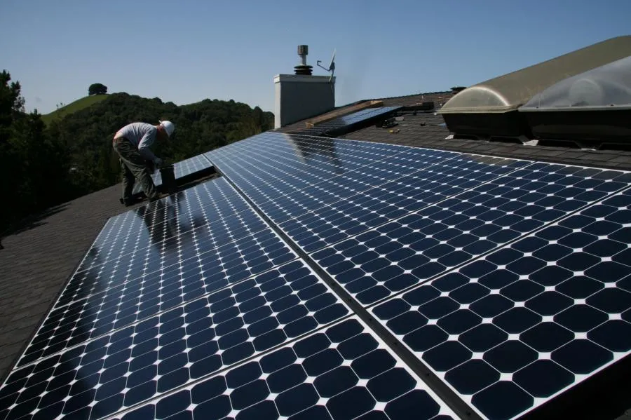  Technician working on solar panel installation on a roof, with tools, clear sky, and surrounding greenery visible.