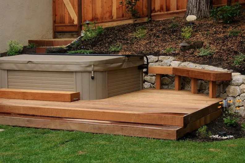  A hot tub is integrated into a wooden deck surrounded by a neatly landscaped garden with a variety of plants and a stone wall.