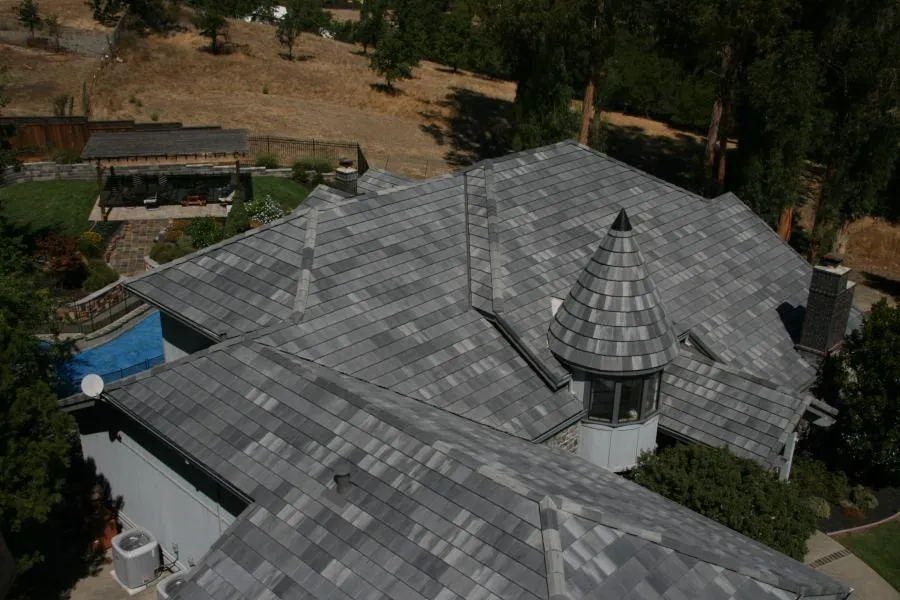  Aerial view of a house with a complex, gray-tiled roof featuring multiple slopes and a turret. Surrounding landscape includes a patio area and greenery.
