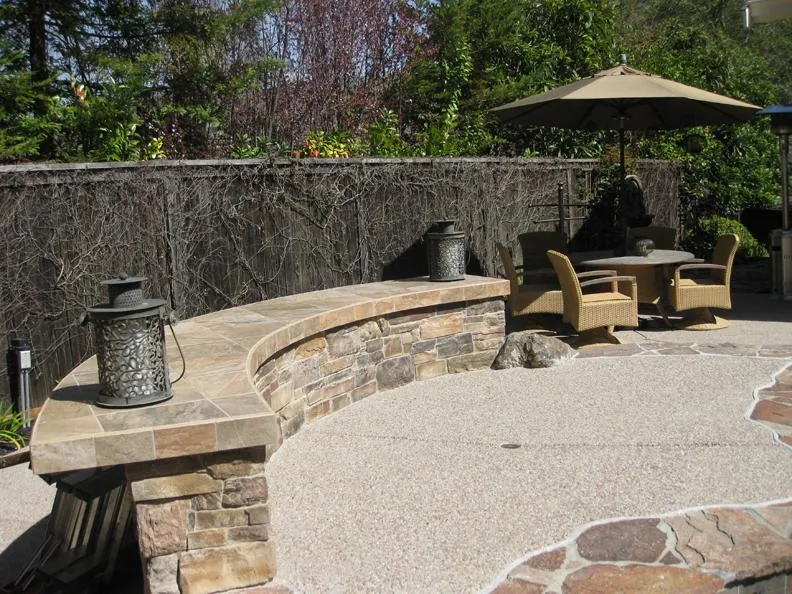 Curved stone bench with lanterns on a patio, featuring a wicker table set under an umbrella, surrounded by a vine-covered fence and greenery.