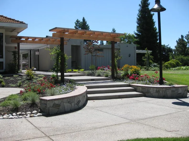  Outdoor area with a pergola over a stone pathway, surrounded by landscaped gardens featuring colorful flowers and shrubs.