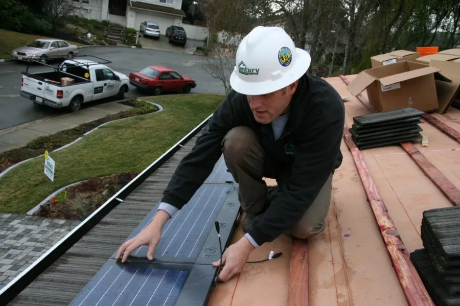  Worker in a hard hat installing solar panels on a roof, with vehicles and houses visible in the background.
