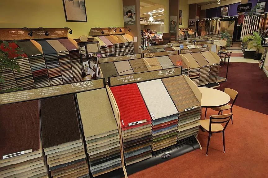 Carpet showroom with multiple displays of colorful carpet samples, a table, chairs, and a warmly lit interior. Carpet showroom with multiple displays of colorful carpet samples, a table, chairs, and a warmly lit interior.
