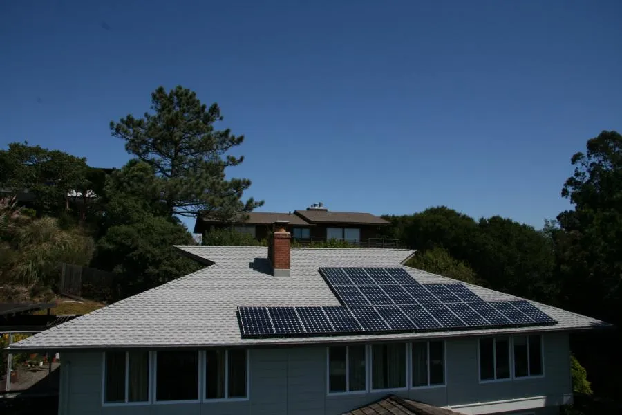  A house with a shingled roof fitted with solar panels, surrounded by trees under a clear blue sky.