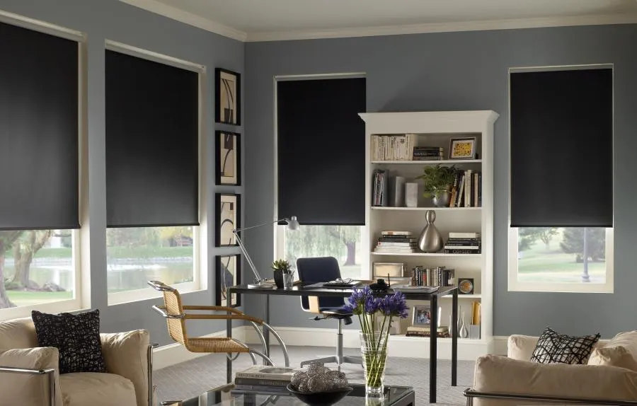  Office with three windows featuring black sunshades, modern desk with chair, bookshelf, artwork on walls, and armchairs.